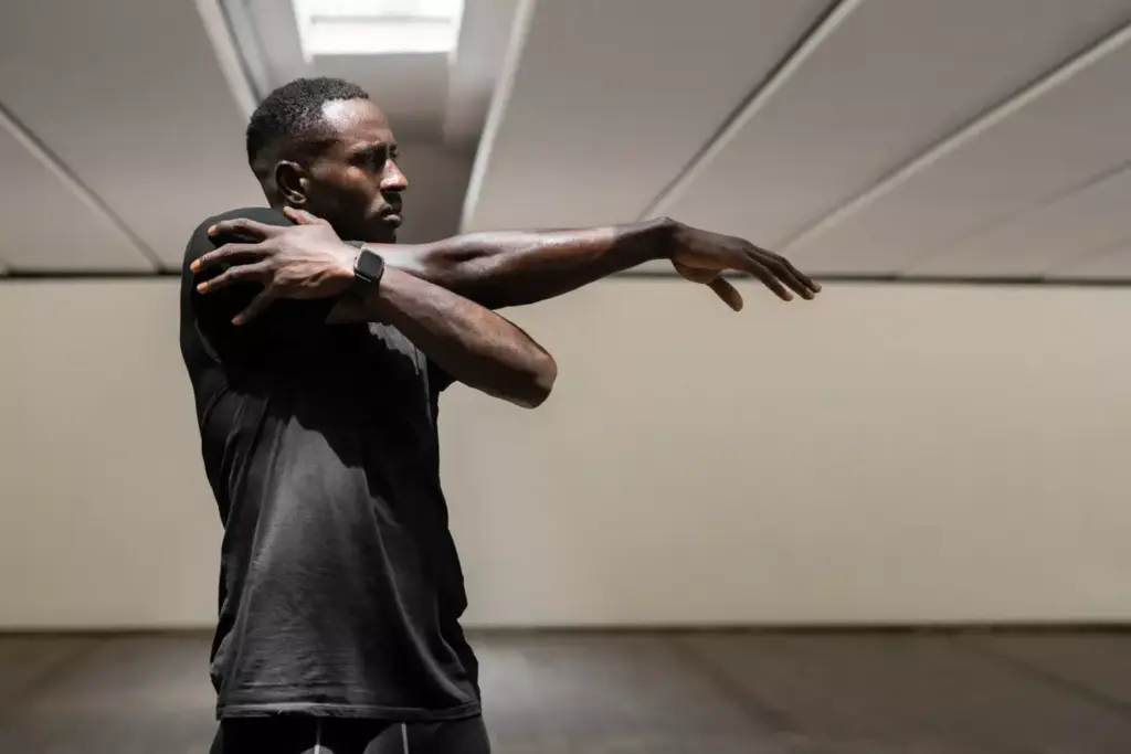 A person performing a shoulder stretch, standing in a dimly lit room with soft, diffused lighting. The subject's face is not visible, but the posture and body language convey a sense of relief and relaxation as they gently pull their arm across their chest, feeling the stretch in their upper back and shoulder. The background is slightly blurred, giving the image a calming, therapeutic atmosphere, suggesting the soothing effects of this exercise on neck and shoulder pain.